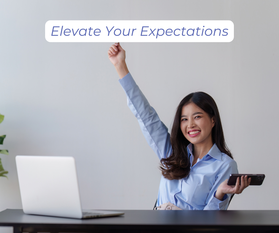 Elevate your expectations: a woman at her desk smiling triumphantly with one arm raised in victory.